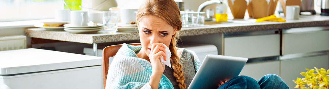 Sick woman sitting in kitchen with tissues and tablet.