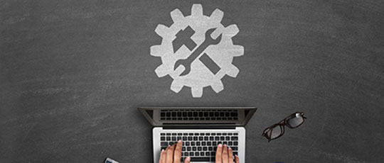 overhead view of hands on laptop keyboard with hammer and wrench criss-crossing on a gear graphic